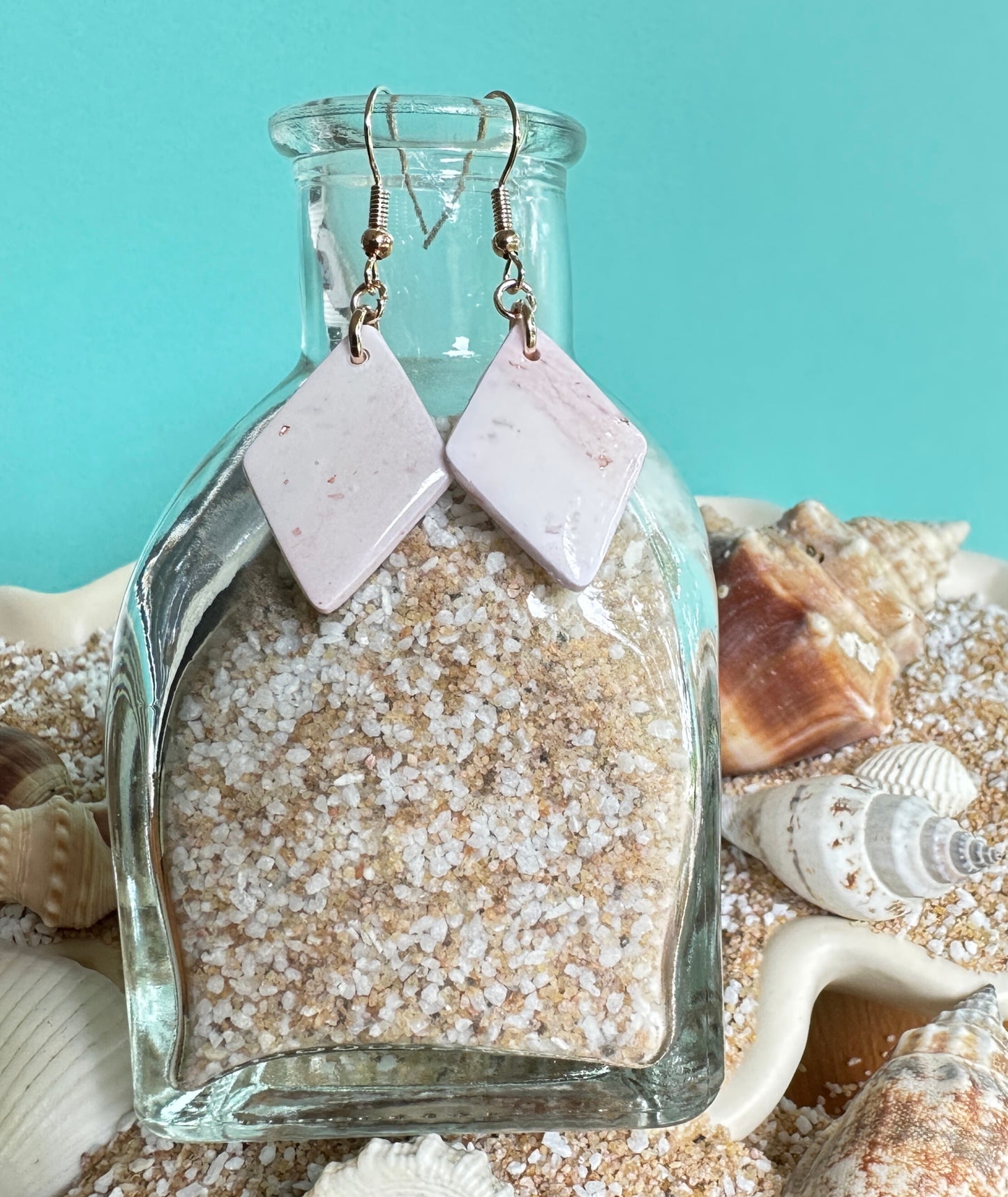 Champagne Shimmer Diamond-Shaped polymer clay dangle earrings hanging on a bottle of sand surrounded by sand and seashells. Colors are light pinks and small gold flecks. Rose gold colored hooks and rings made of  nickel-free iron.