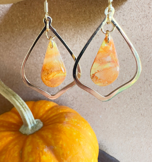 Harvest Celebration polymer clay dangle earrings. Teardrop-shaped center piece with pear-shaped gold colored hardware. Colors of orange and yellow marbled together with gold shimmer and gold foil flakes. Gold colored hooks and rings. Pumpkin in the background.
