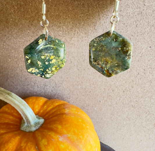 Enchanted Moss polymer clay dangle earrings. Hexagon-shaped. Colors of greens, blues, and browns marbled together with gold foil flakes. Gold colored hooks and rings. Pumpkin in the background.