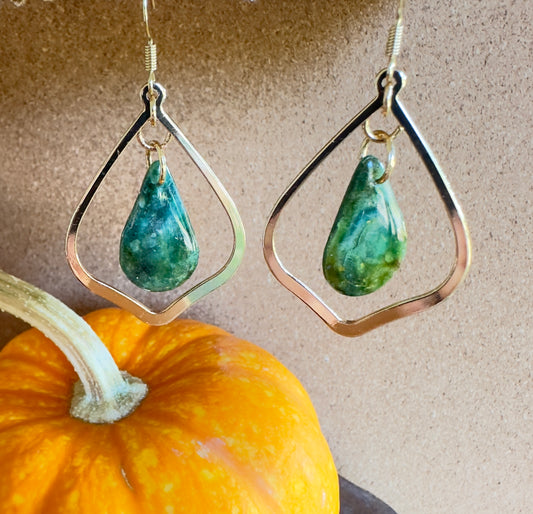 Enchanted Moss polymer clay dangle earrings. Teardrop-shaped center piece with pear-shaped gold colored hardware. greens, blues, and browns marbled together with gold foil flakes. Gold colored hooks and rings. Pumpkin in the background.
