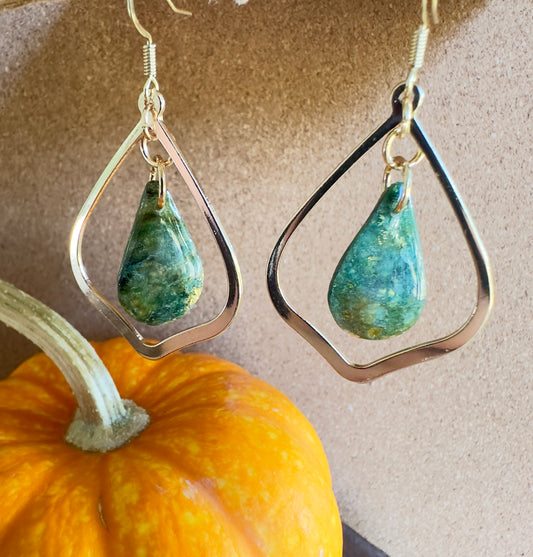 Enchanted Moss polymer clay dangle earrings. Teardrop-shaped center piece with pear-shaped gold colored hardware. greens, blues, and browns marbled together with gold foil flakes. Gold colored hooks and rings. Pumpkin in the background.