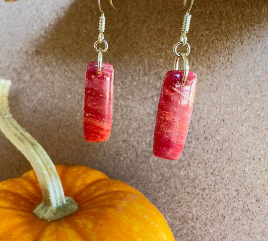 Macintosh polymer clay dangle earrings. Vertical rectangle shape with colors of red, cranberry, gold shimmer, and gold foil flakes. Gold colored hooks and rings. Pumpkin in the background.