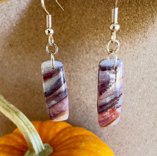 Autumn Vinyard polymer clay dangle earrings. Vertical rectangle shape with colors of translucent white, purples, mauves, and rose gold foil flakes. Rose Gold colored hooks and rings. Pumpkin in the background.