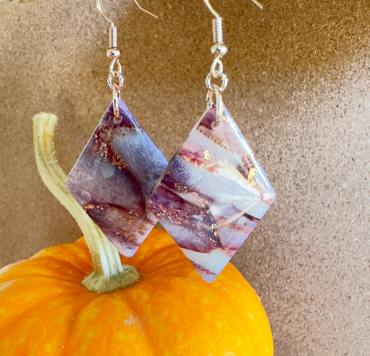 Autumn Vinyard polymer clay dangle earrings. Diamond Shape with colors of translucent white, purples, mauves, and rose gold foil flakes. Rose Gold colored hooks and rings. Pumpkin in the background.