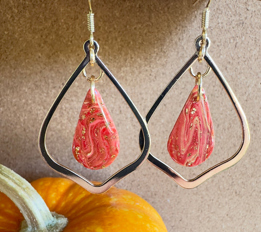 Orchard Swirl polymer clay dangle earrings. Teardrop-shaped center piece with pear-shaped gold colored hardware. Colors of red and yellow swirled together with gold foil flakes. Gold colored hooks and rings. Pumpkin in the background.
