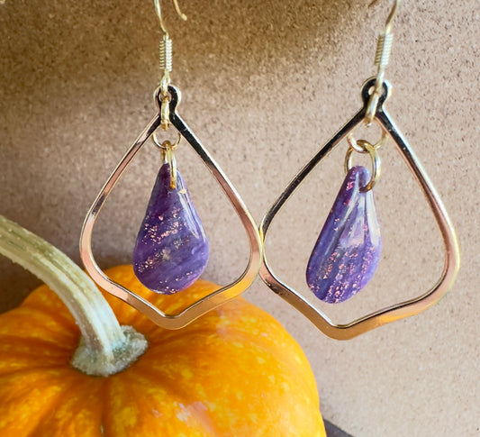 Witch's Brew polymer clay dangle earrings. Teardrop-shaped center piece with pear-shaped gold colored hardware. Colors of purple swirled together with rose gold foil flakes. Gold colored hooks and rings. Pumpkin in the background.