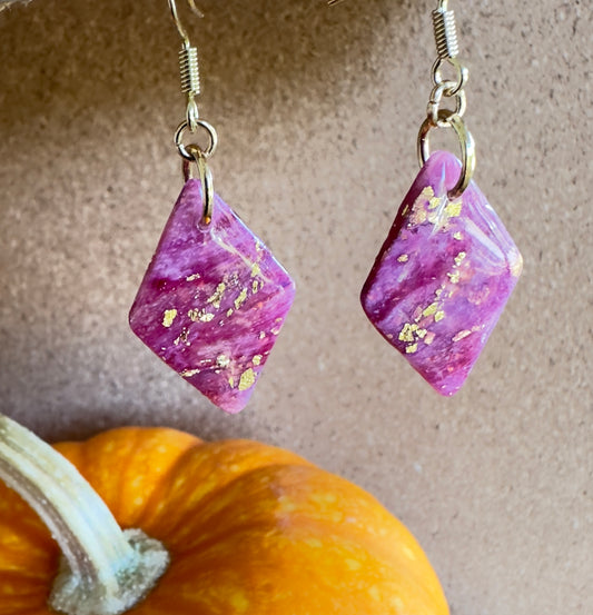 Cranberry Mimosa diamond-shaped polymer clay dangle earrings. Diamond-shaped. Coiors of fuschia, purple, translucent white, and gold foil flakes. Gold colored hooks and rings. Pumpkin in the background.