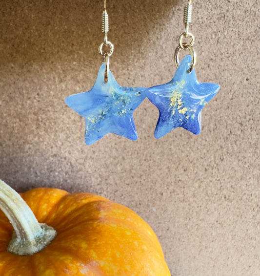 Blue Moon Star-shaped dangle polymer clay earrings. Colors of translucent white, light and medium blues, with gold foil flakes. Gold colored hooks and rings. Pumpkin in the background.